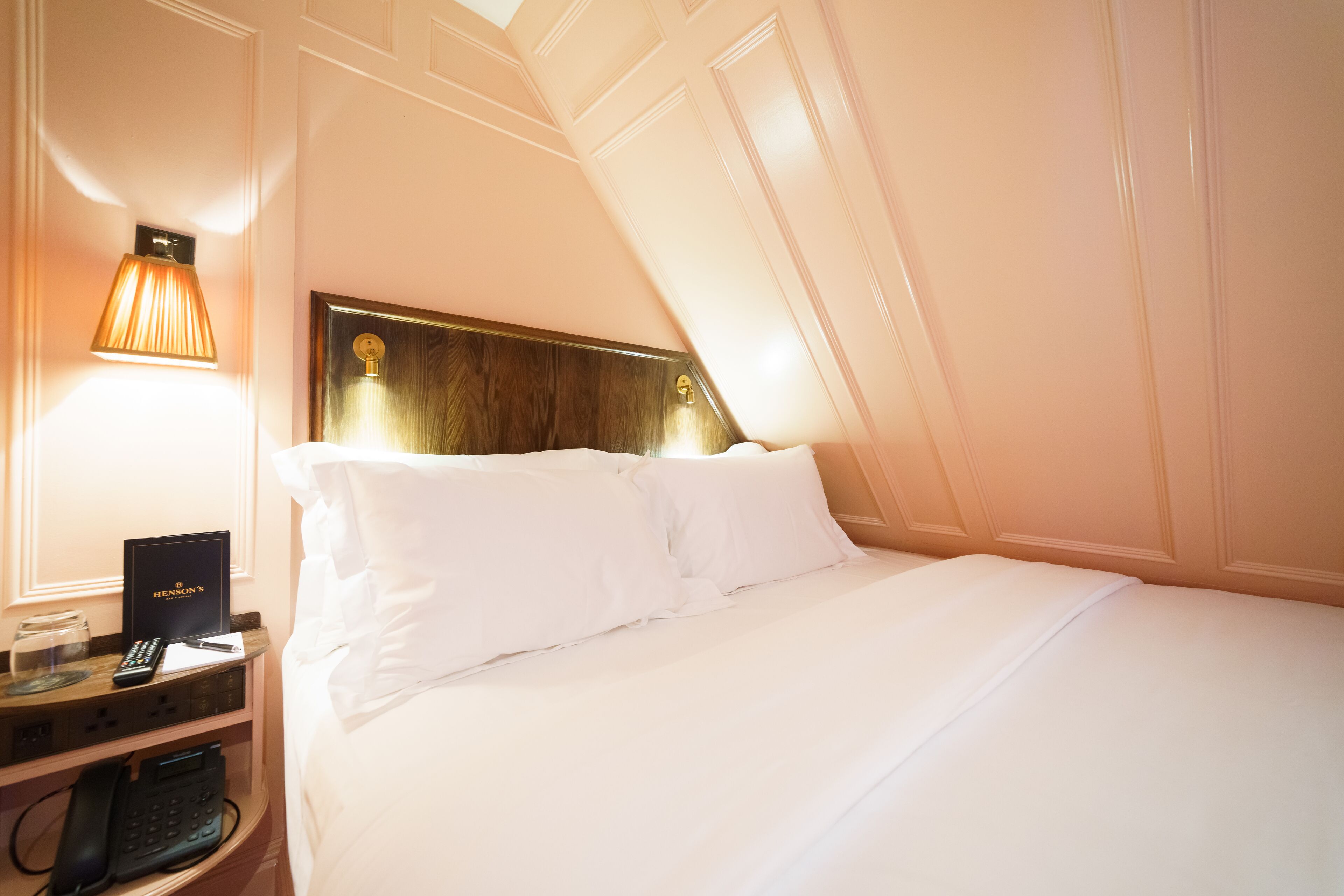A hotel room with a neatly made bed, a bedside table with a lamp, a remote control, a book, and a phone.