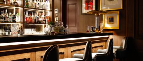A bar area with a wooden bar counter, multiple liquor bottles, glasses, and two leather bar stools.