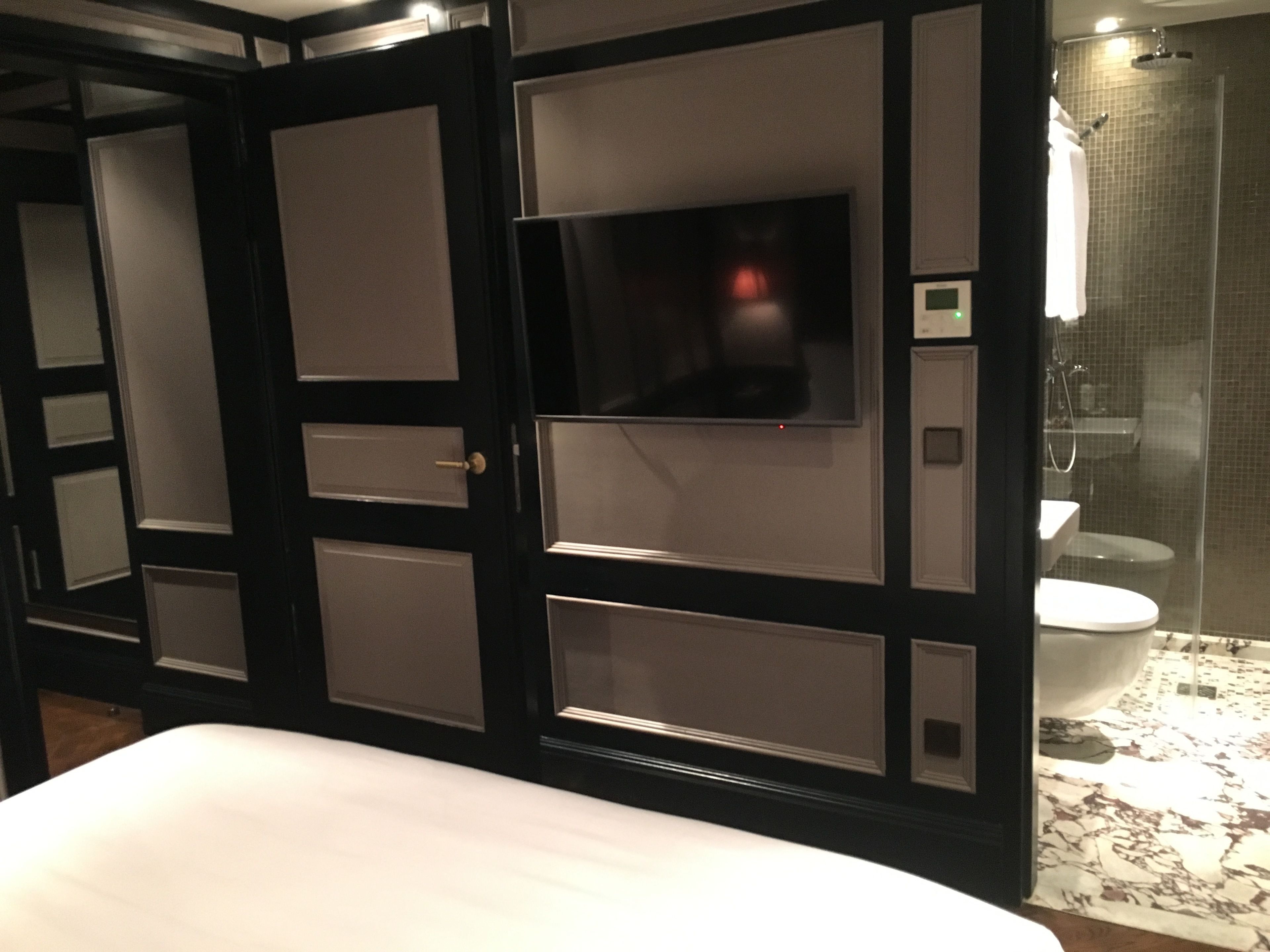 A hotel room with a bed, a television mounted on a dark panel wall, a shower area, and a toilet.