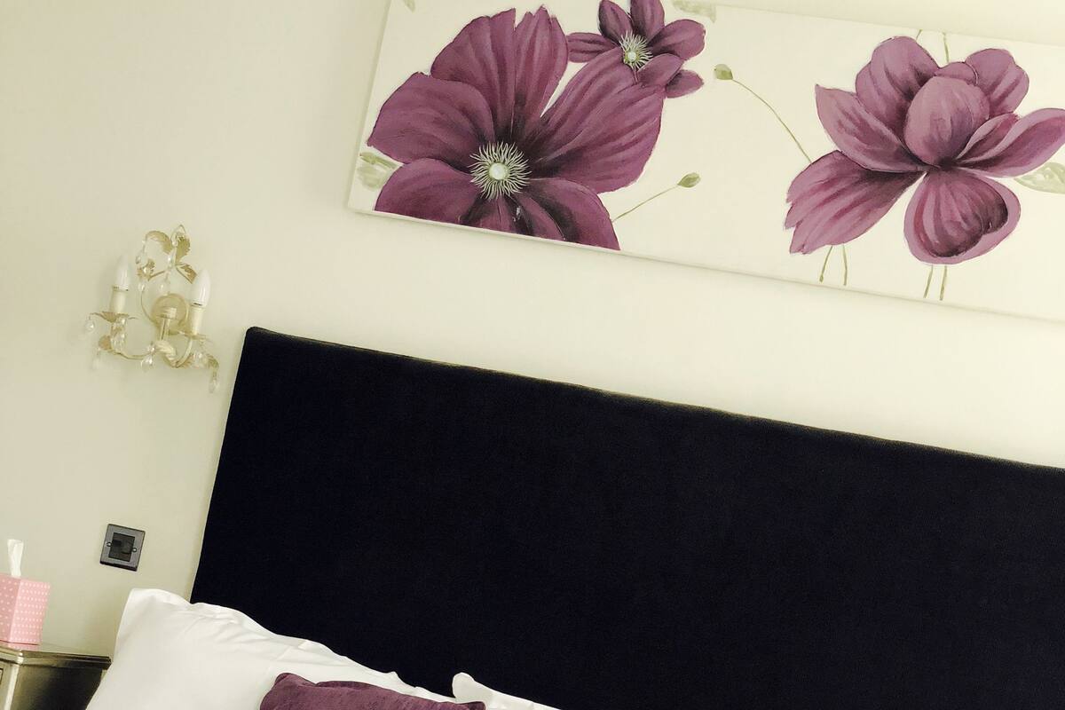 Un lit bien fait, avec une literie blanche et violette, une tête de lit sombre et une œuvre d’art florale accrochée au mur.