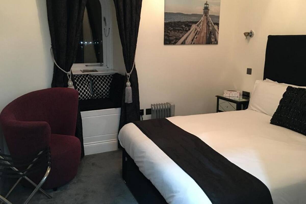 Une chambre d’hôtel avec un lit, une chaise rouge, une fenêtre avec des rideaux, un radiateur et un tableau représentant un phare.