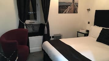 A hotel room with a bed, a red chair, a window with curtains, a radiator, and a painting of a lighthouse.