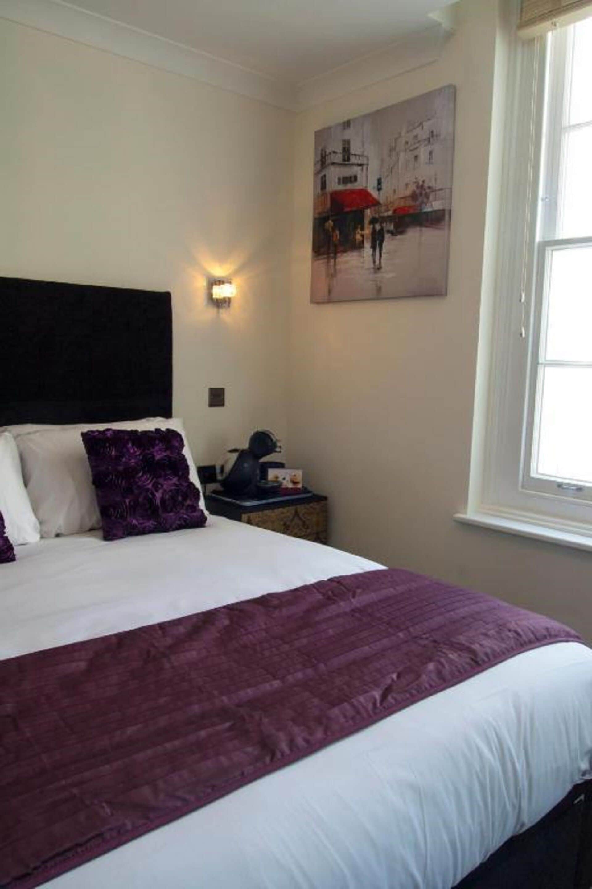 A neatly made bed with a white and purple bedspread, a black headboard, and a wall-mounted light fixture.
