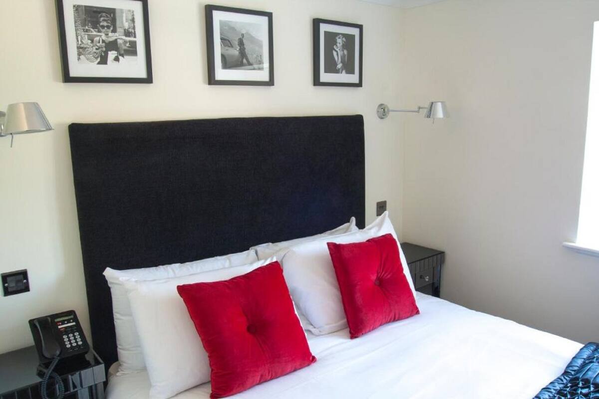 Une chambre d’hôtel avec un grand lit, du linge de lit blanc, deux oreillers rouges, une tête de lit noire, une table de chevet avec une lampe, un téléphone et trois tableaux encadrés accrochés au mur.