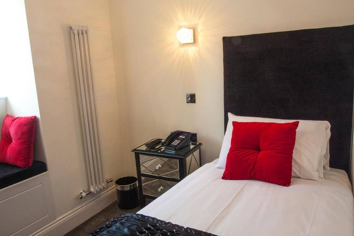 Une chambre d’hôtel avec un lit, une table de chevet avec un téléphone, un radiateur et une fenêtre avec un rideau.