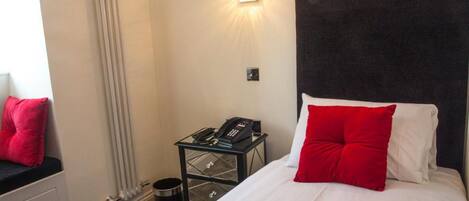 A hotel room with a bed, a nightstand with a phone, a radiator, and a window with a curtain.