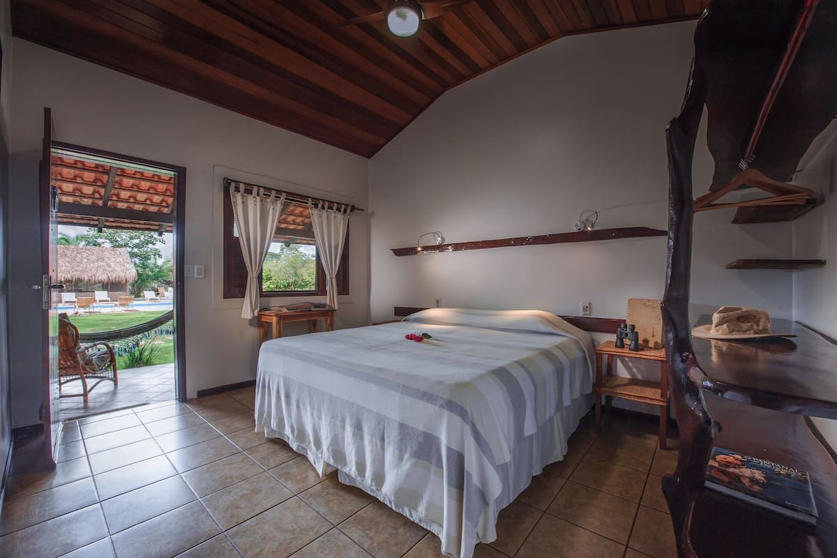 Un dormitorio con cama, techo de madera, ventana con cortinas, una mesita con lámpara y vista al exterior.