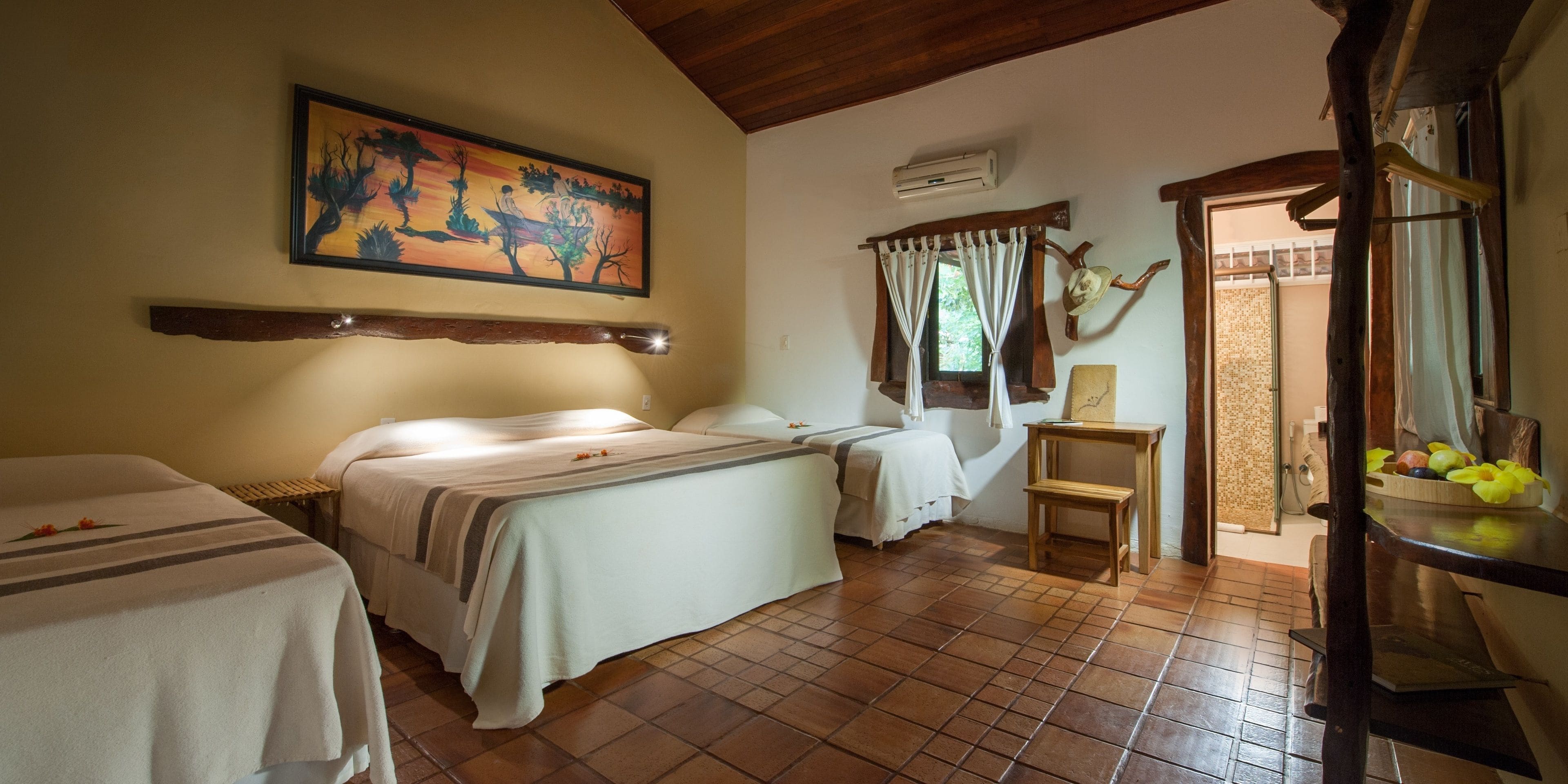 Una habitación con dos camas, un cuadro en la pared, una ventana con cortinas, una silla de madera y una mesa con frutas.