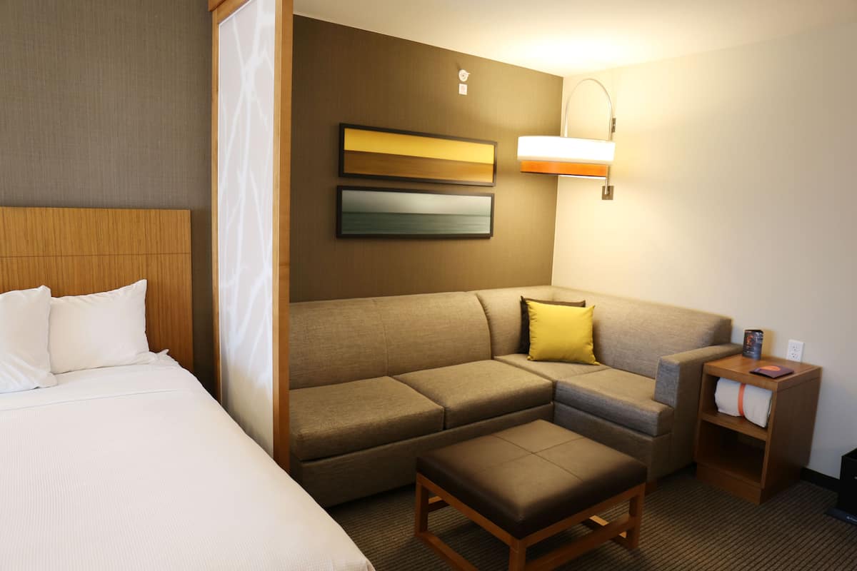Habitación de hotel con cama, sofá, reposapiés, mesita de noche y lámpara de pared.