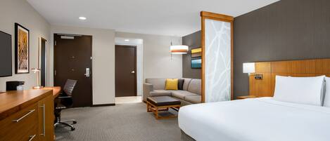A hotel room with a large bed, a desk with drawers, a sofa with cushions, a small table, and a wall-mounted TV.