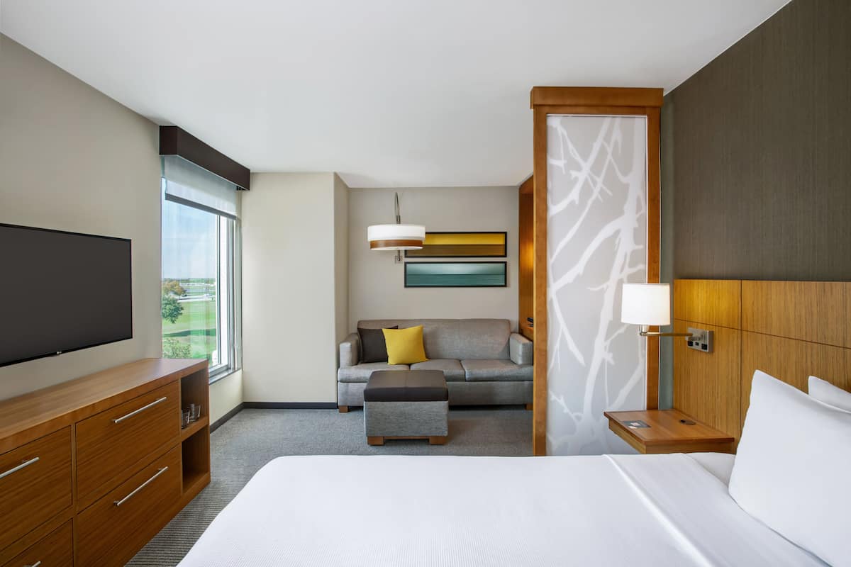 Habitación de hotel con una cama grande, un sofá, un televisor, una mesita de noche de madera y una ventana con vistas.