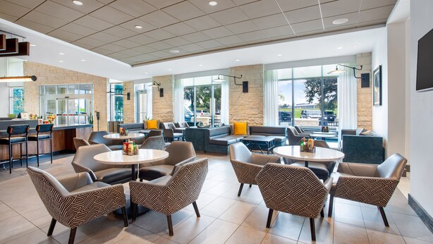 A modern indoor seating area with round tables and patterned chairs, a kitchen counter, and large windows offering a view of the outdoors. - Hyatt Place Austin Airport (Austin)