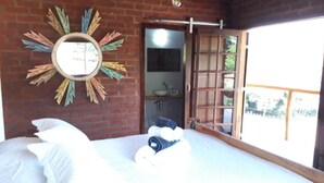 A room with a bed, a mirror with a colorful design, a wooden door, and a view of greenery outside.