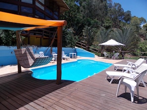Um deck de madeira com piscina, cadeiras de descanso brancas, uma rede e uma área de estar coberta.