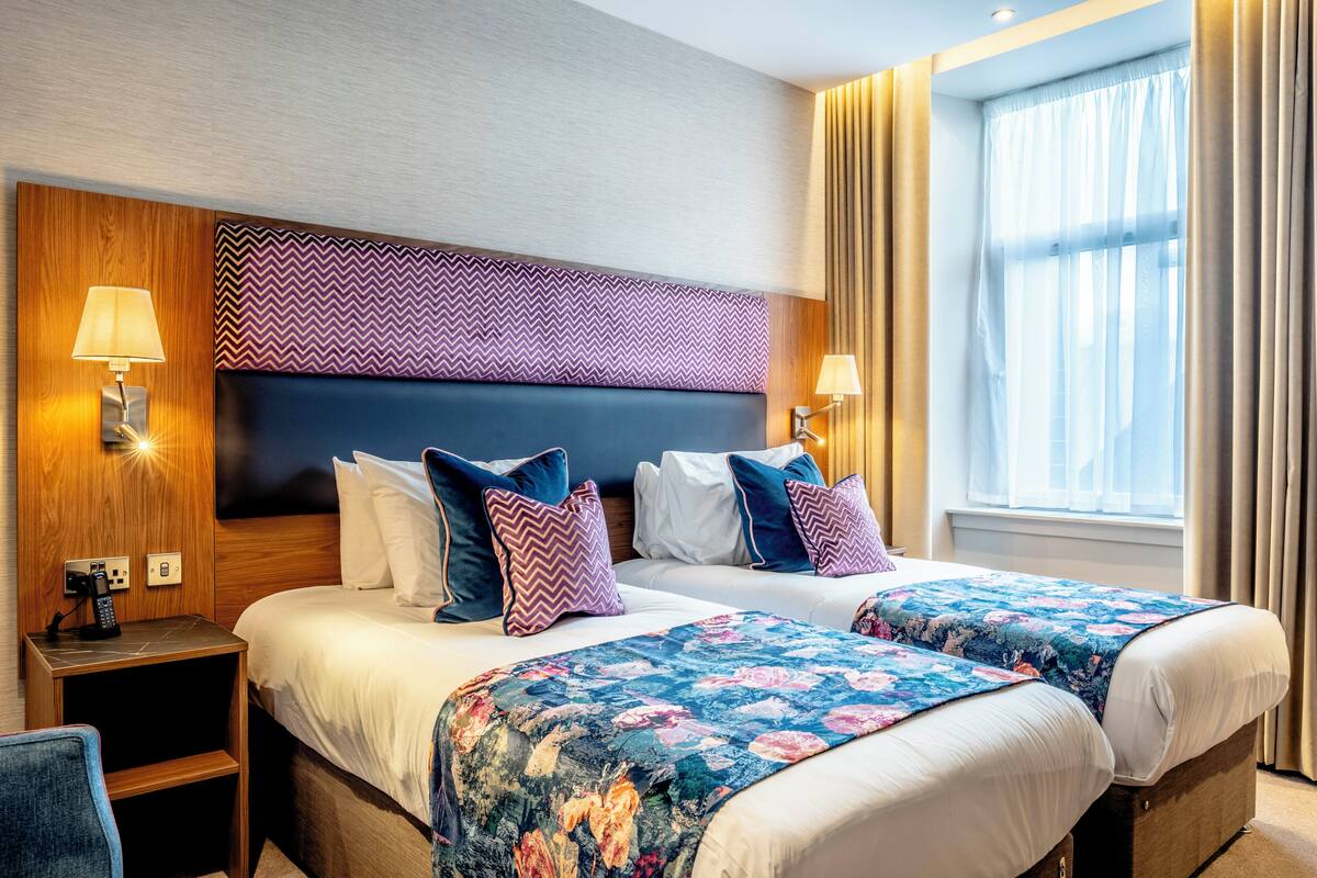 A hotel room with two beds, a wooden headboard, patterned cushions, bedside lamps, and a window with curtains.
