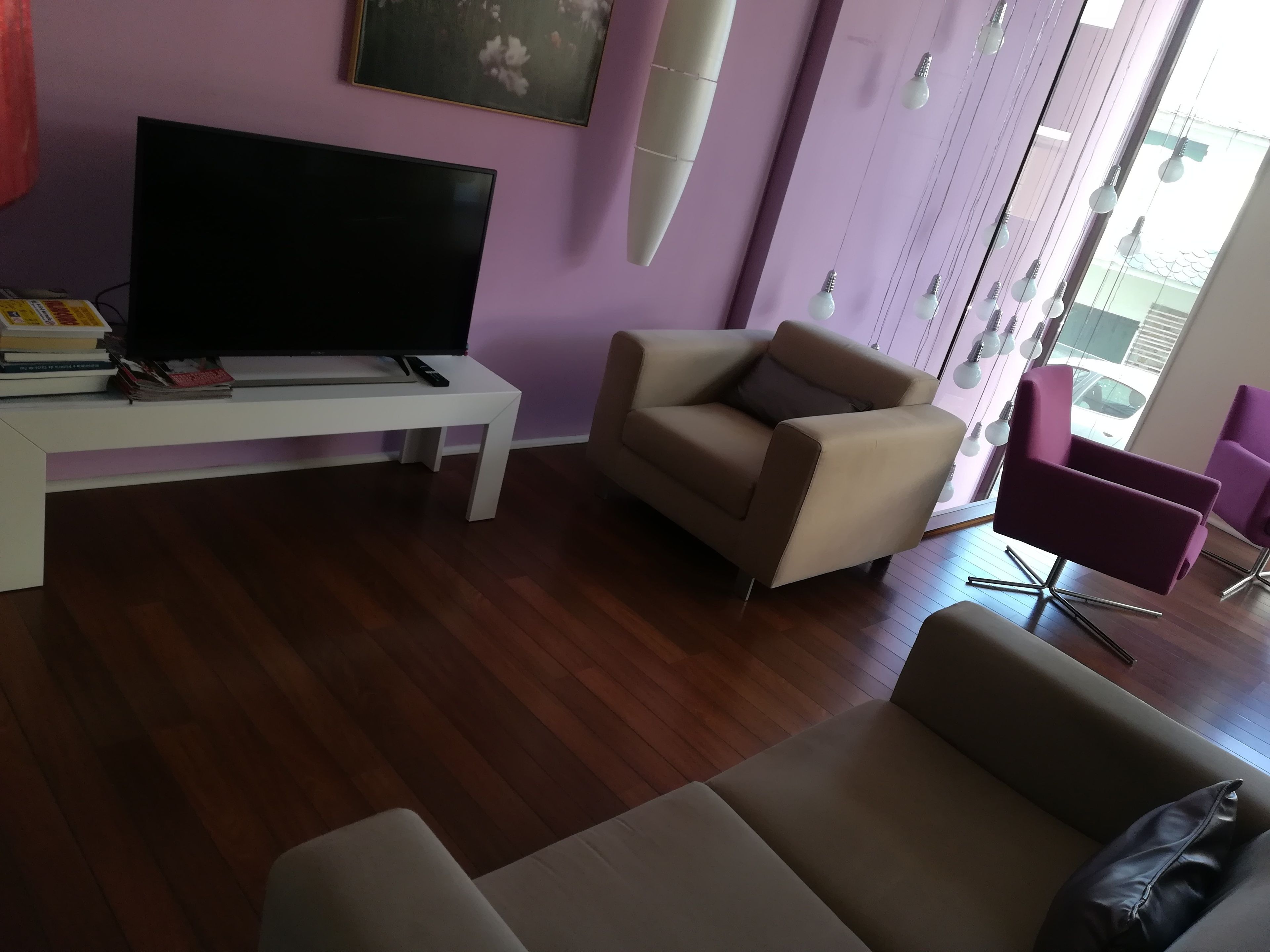 Una sala de estar con un televisor de pantalla plana sobre una mesa blanca, un sofá color beige y un sillón púrpura.
