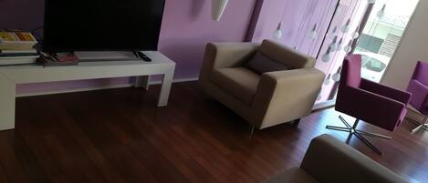 Una sala de estar con un televisor de pantalla plana sobre una mesa blanca, un sofá color beige y un sillón púrpura.