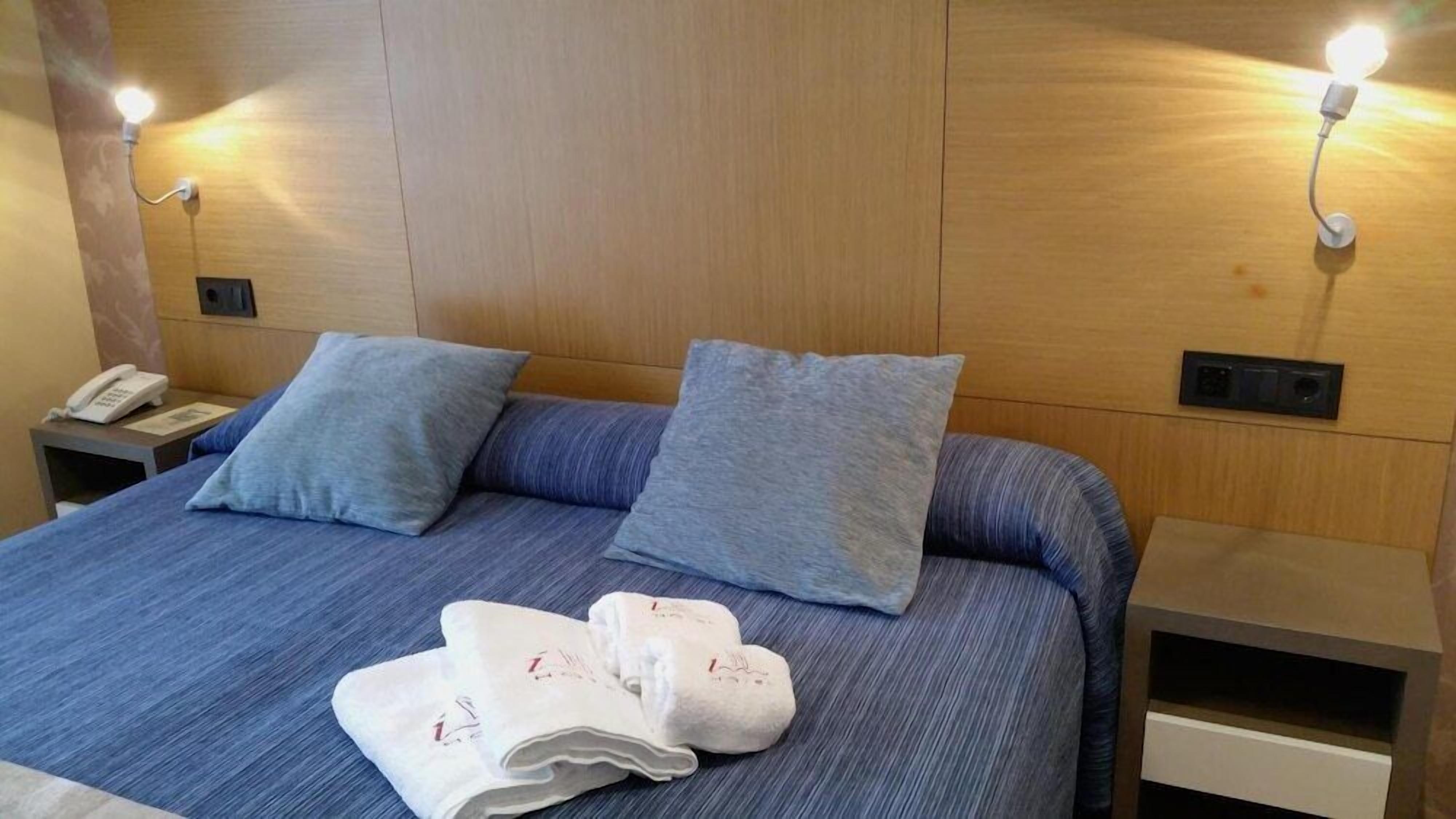 A hotel room with a bed, two pillows, a bedside table with a phone, and a wall-mounted light fixture.