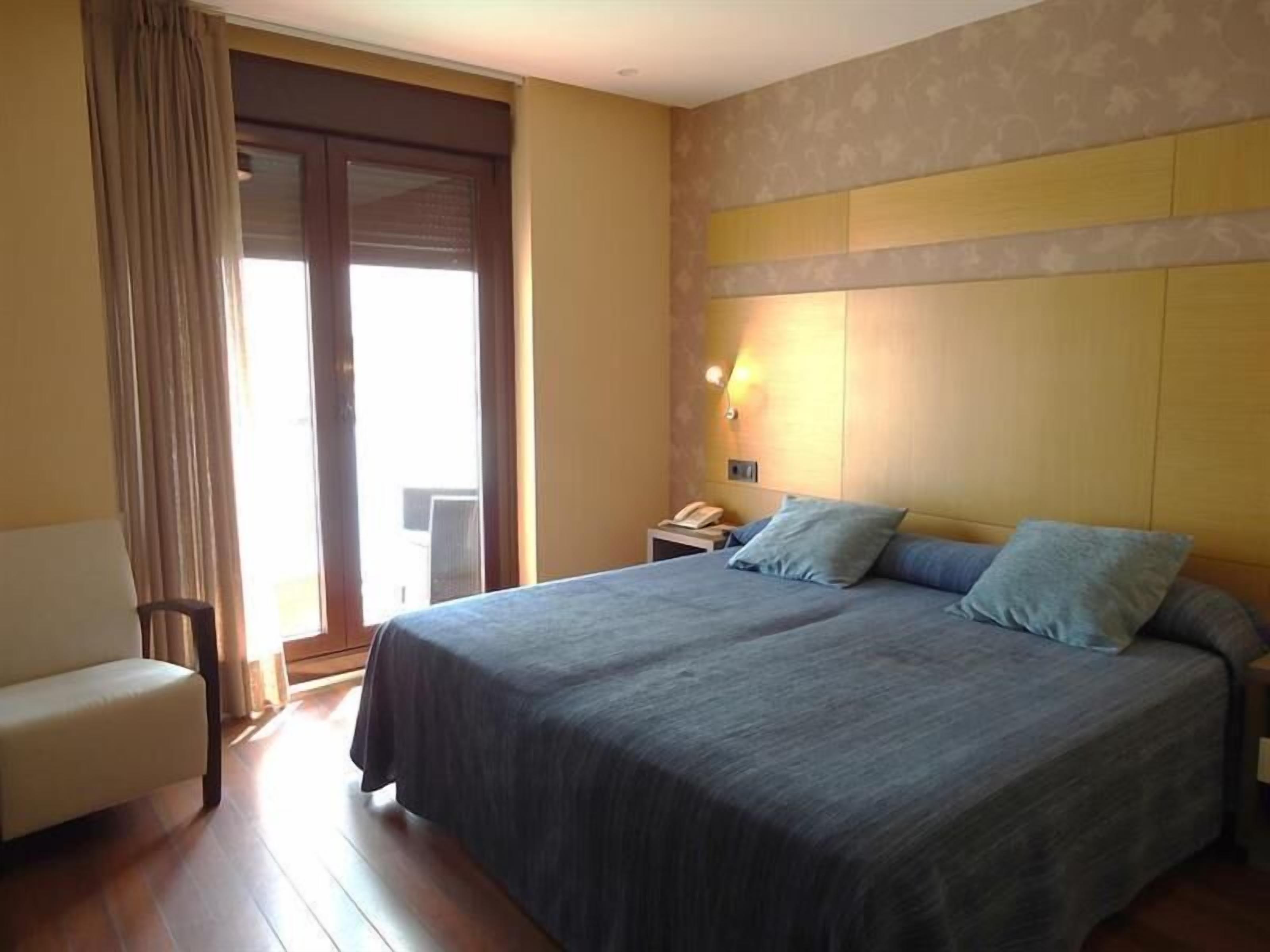 Una habitación de hotel con una cama, una silla, una mesita de noche con un teléfono y una ventana con cortinas.