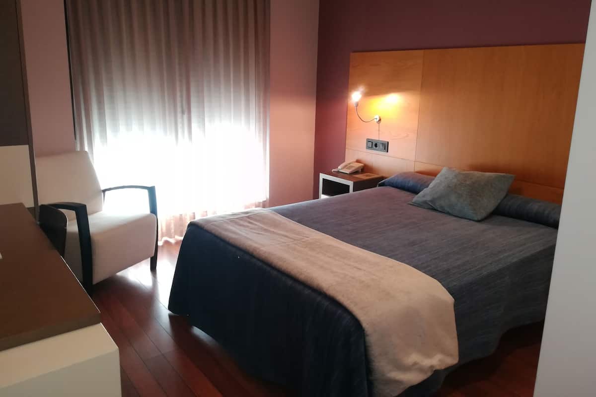 A hotel room with a bed, a desk, a chair, a nightstand with a phone, and a window with curtains.