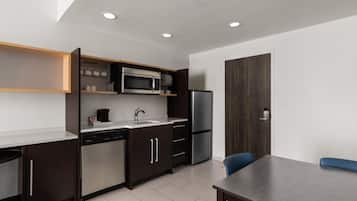 A modern kitchen with a built-in microwave, refrigerator, and dishwasher. There is a dining table with chairs and a wooden cabinet with shelves above the counter.