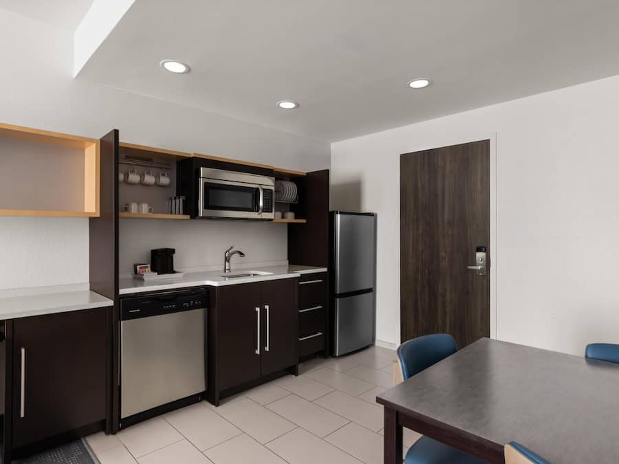A modern kitchen with a built-in microwave, refrigerator, and dishwasher. There is a dining table with chairs and a wooden cabinet with shelves above the counter.
