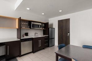 Una cocina moderna con microondas integrado, refrigerador y lavavajillas. Hay una mesa de comedor con sillas y un mueble de madera con estantes sobre la encimera.