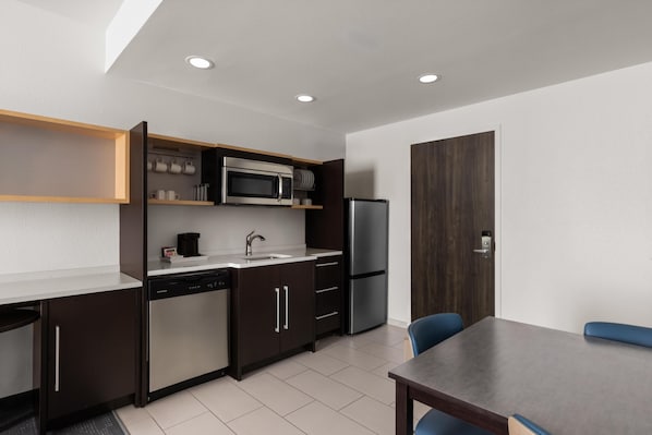 A modern kitchen with a built-in microwave, refrigerator, and dishwasher. There is a dining table with chairs and a wooden cabinet with shelves above the counter. - Home2 Suites by Hilton Gonzales, LA (Gonzales)