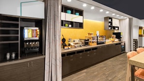 Un bar à petit-déjeuner moderne, équipé d’une machine à café, d’un présentoir de fruits et d’un espace détente avec des chaises orange.