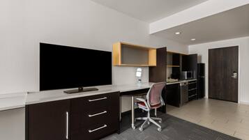 A modern office space with a large flat-screen TV, a desk, a chair, a microwave, and a refrigerator.