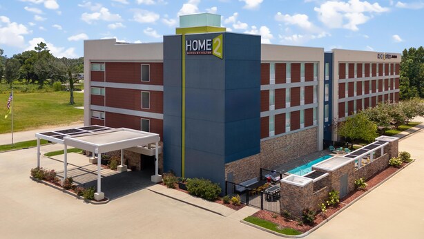 Un bâtiment d’hôtel avec une enseigne indiquant « HOME 2 Suites by Hilton ». Il y a un espace extérieur couvert avec une piscine et des chaises longues.