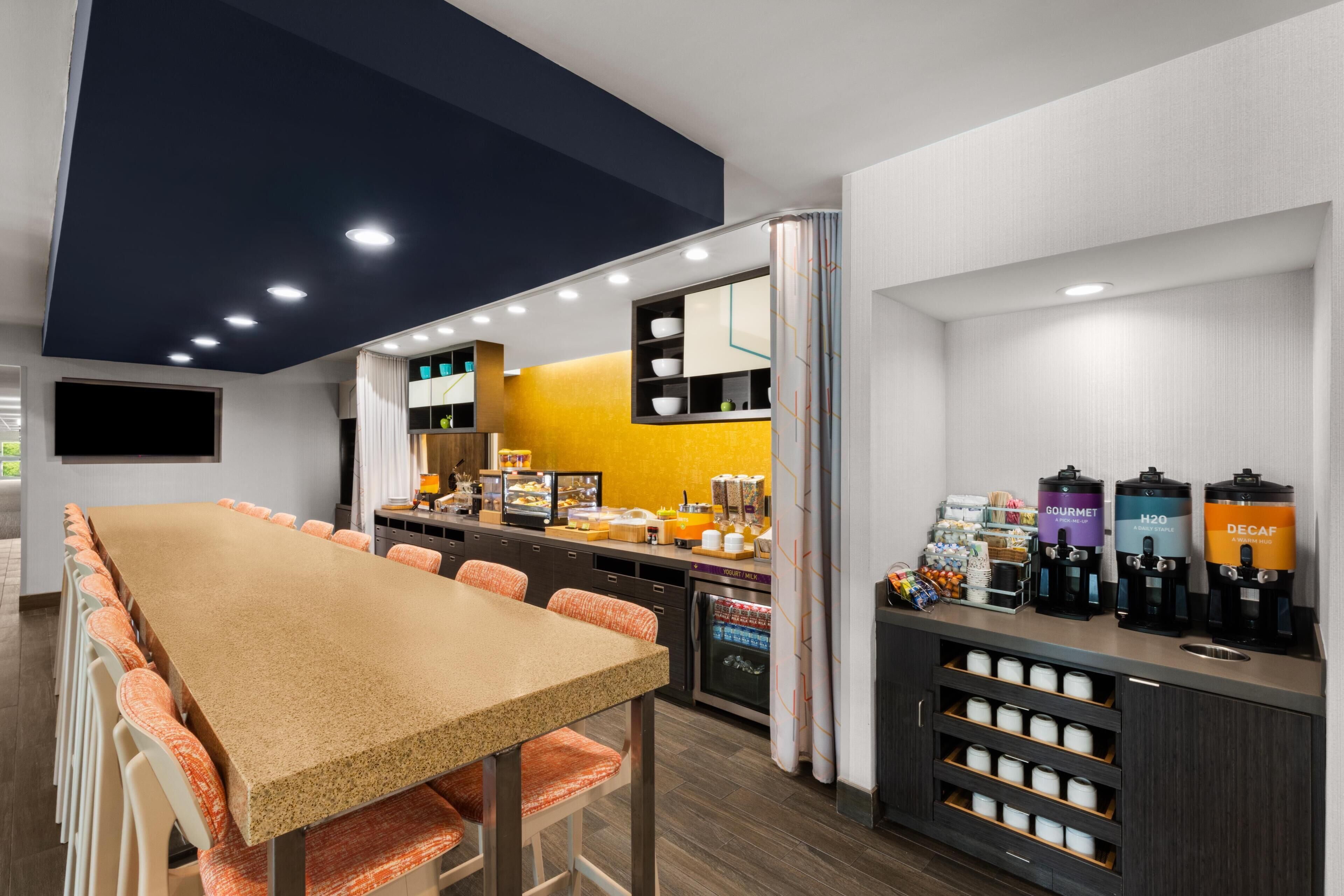 A modern cafe with a long table, chairs, a counter with coffee machines, and a refrigerator.