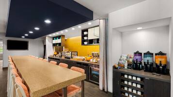 A modern cafe with a long table, chairs, a counter with coffee machines, and a refrigerator.