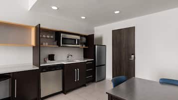 A modern kitchen with a built-in microwave, refrigerator, and dishwasher. There is a dining table with chairs and a wooden cabinet with shelves above the counter.
