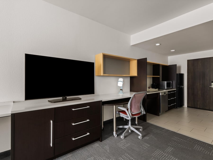 A modern office space with a large flat-screen TV, a desk, a chair, a microwave, and a refrigerator.