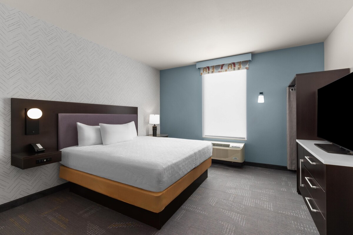 Een hotelkamer met een bed, nachtkastje, lamp, raam, airconditioning en een televisie.