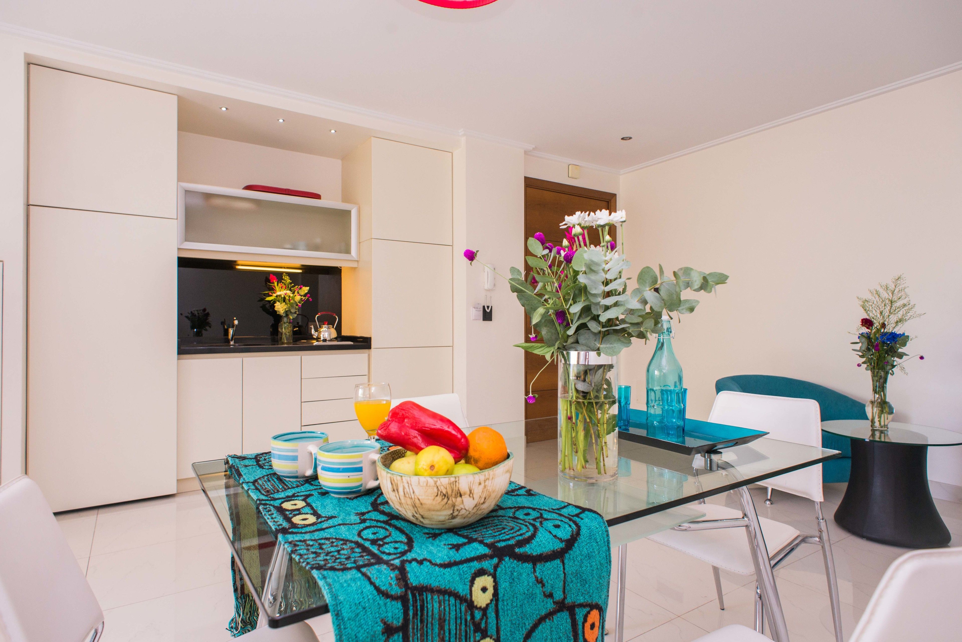 A modern kitchen with white cabinets, a glass table with a teal tablecloth, and a bowl of fruit.