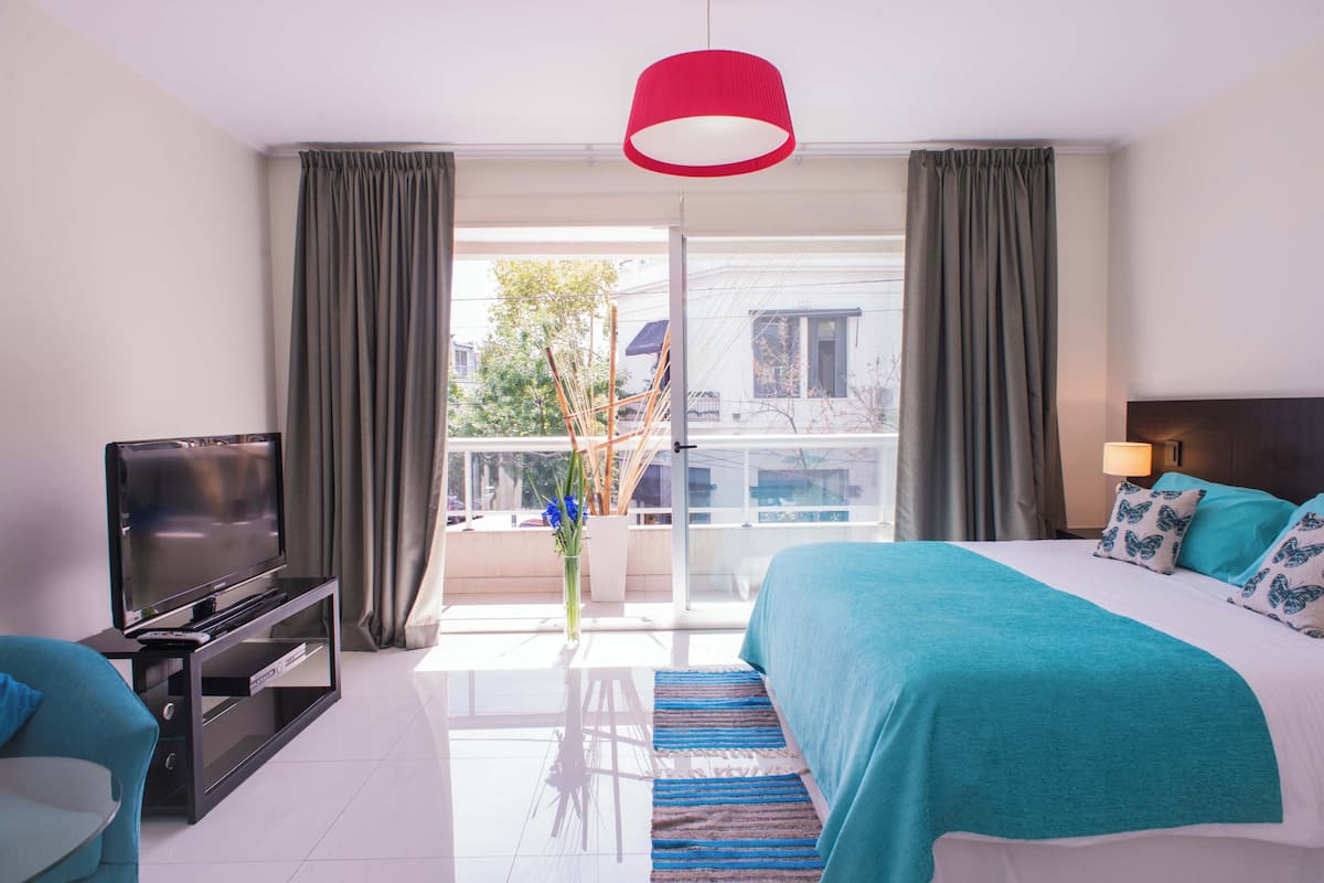 Habitación de hotel con cama, televisión, balcón con cortinas y vistas a edificios.
