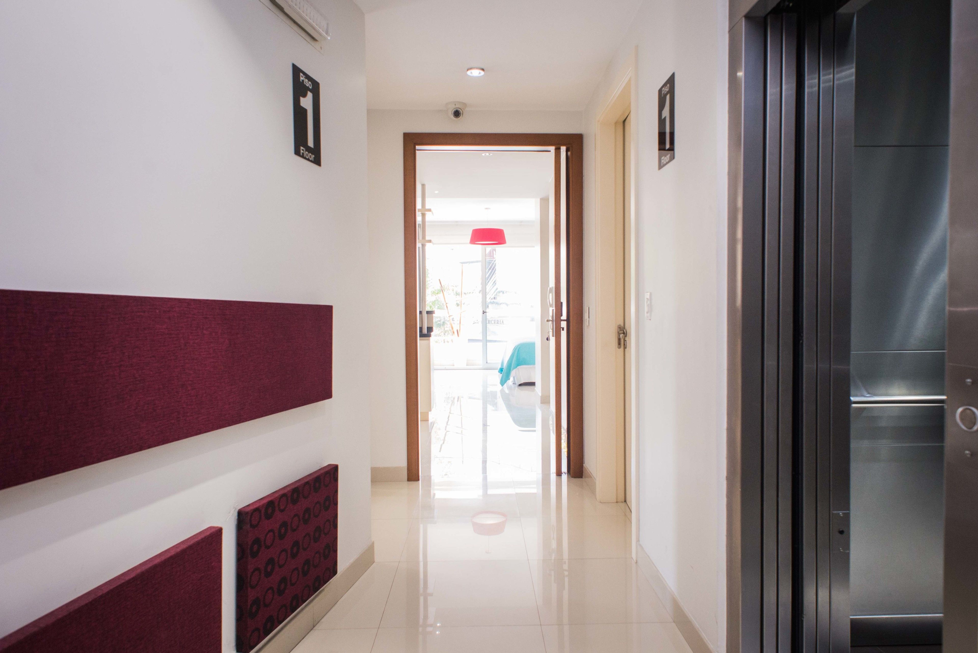 A hallway with a red padded wall, a glass door leading to a room, and a metallic elevator.