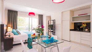 Ein modernes Hotelzimmer mit Bett, Esstisch mit Stühlen, Küchenzeile und großen Fenstern mit Vorhängen.