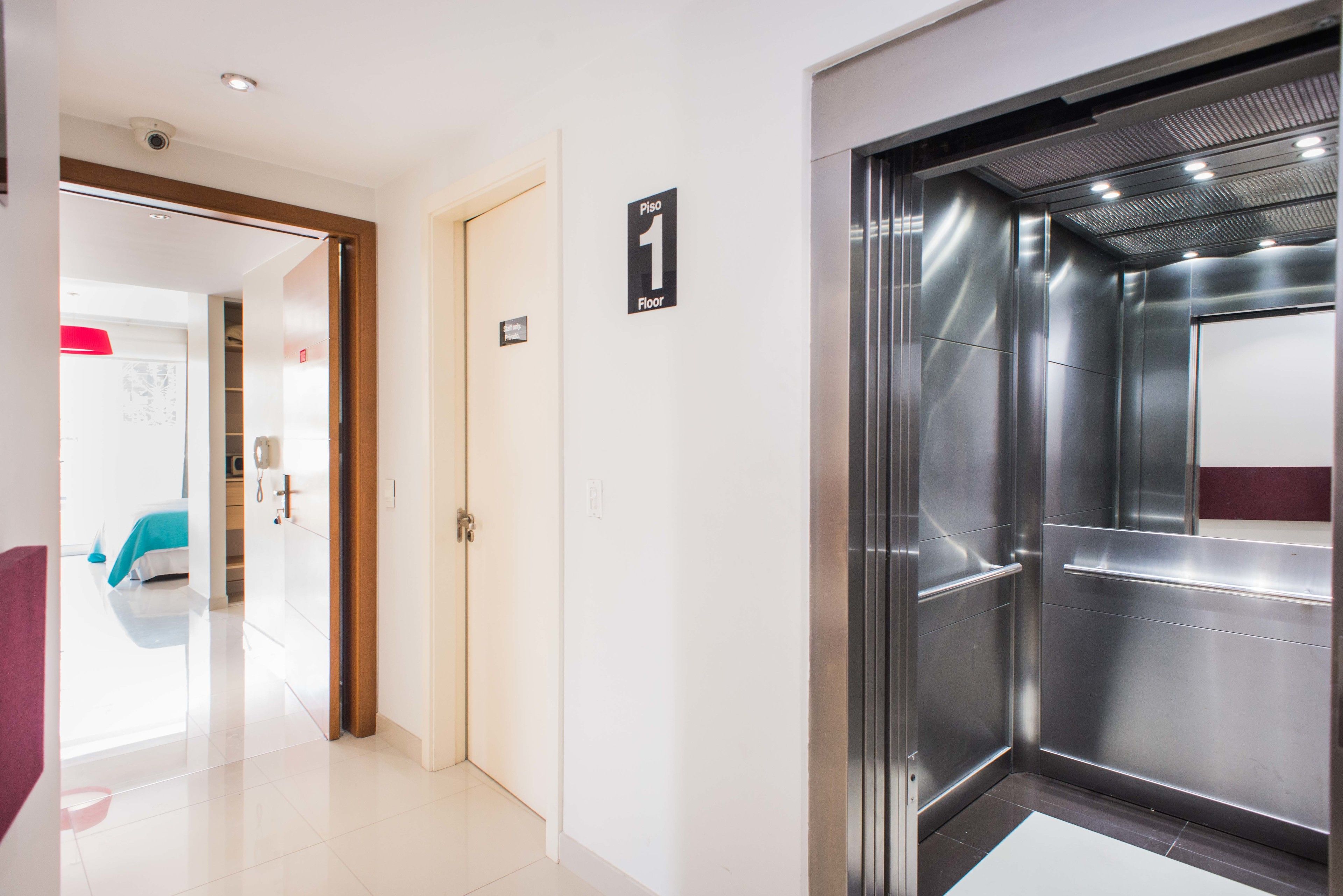 A modern elevator with a metallic finish, a visible floor indicator, and a clean, well-lit hallway leading to a room with a bed and a red chair.