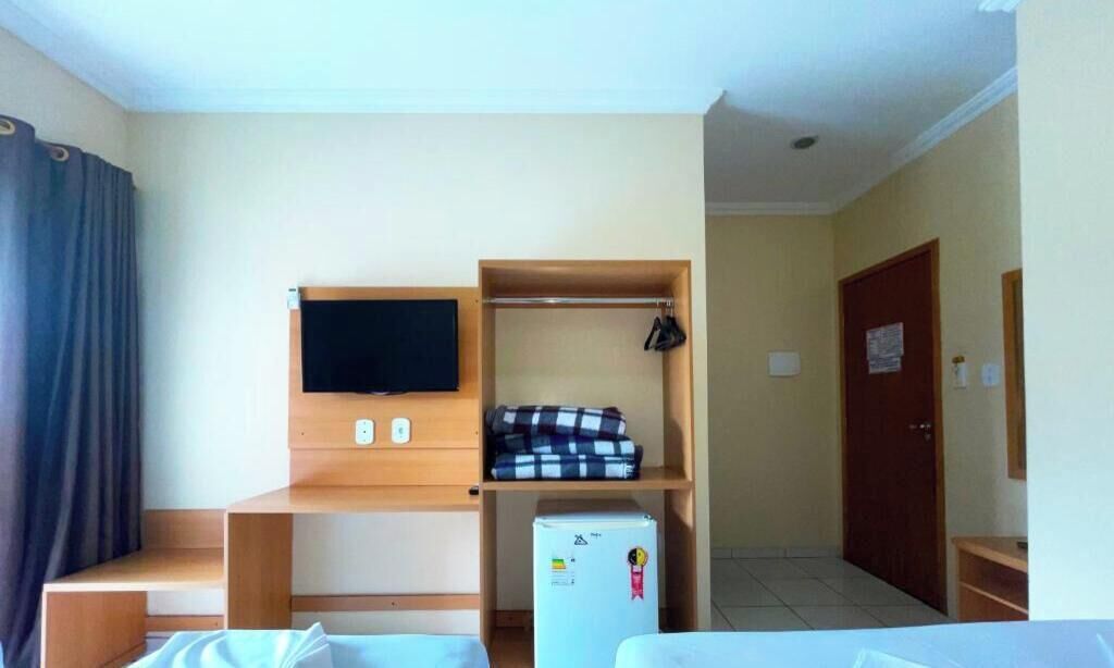 A hotel room with a bed, a TV mounted on the wall, a small refrigerator, and a wooden shelf with folded clothes.