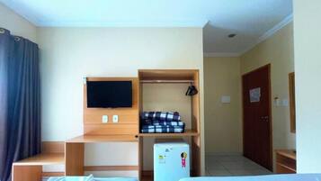 A hotel room with a bed, a TV mounted on the wall, a small refrigerator, and a wooden shelf with folded clothes.