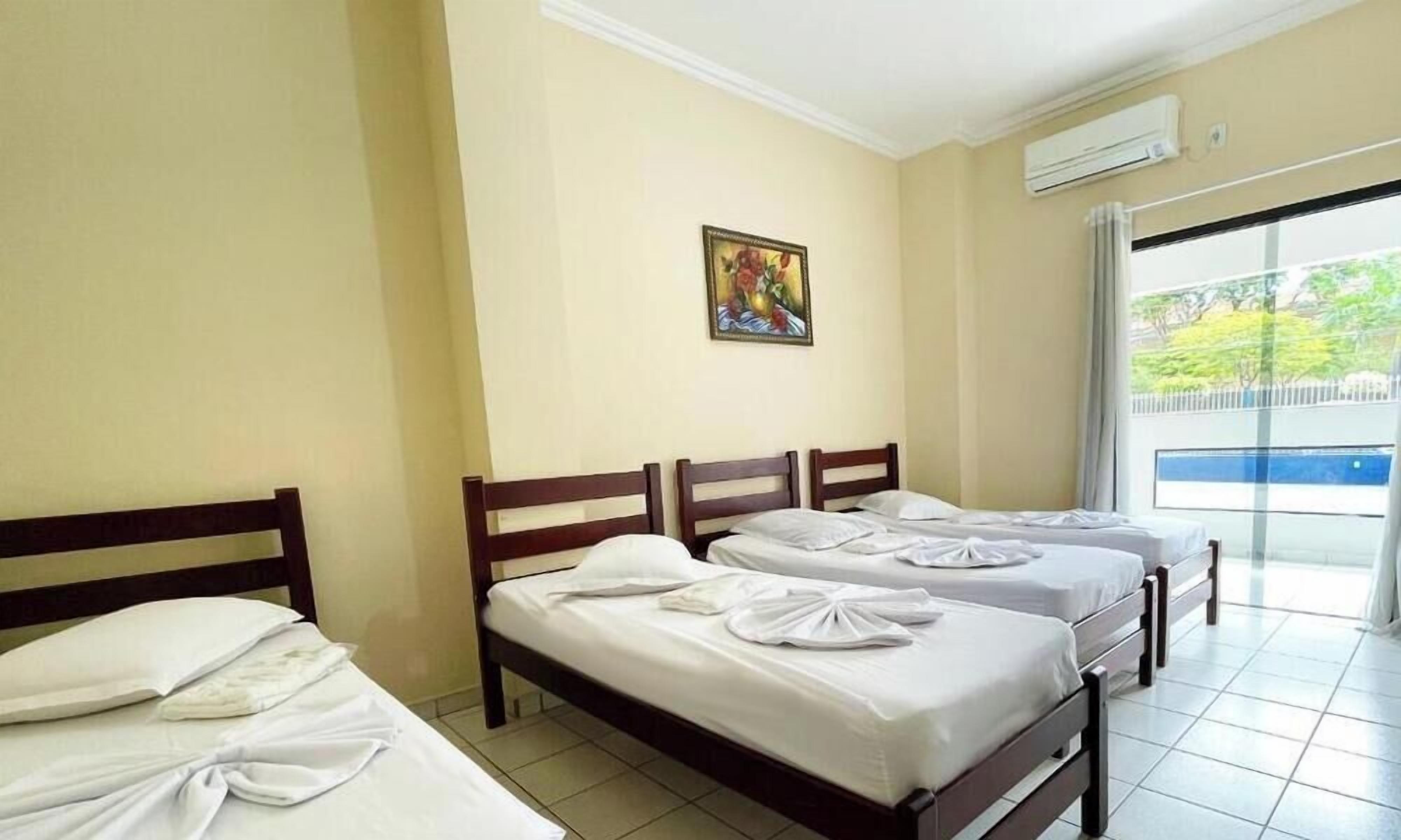 A hotel room with three beds, a painting on the wall, and a view of the outdoors through a large window.