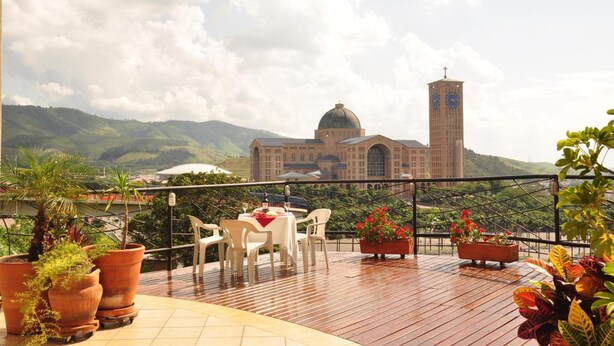 Une terrasse avec une table dressée pour deux personnes, offrant une vue sur un grand bâtiment doté d’un dôme et d’une tour horloge, le tout entouré de verdure et de collines.