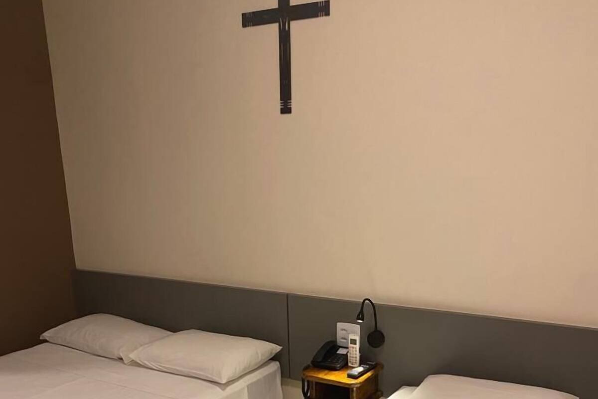 A hotel room with two single beds, a nightstand with a phone and lamp, and a cross on the wall.