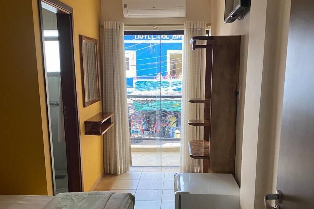 A hotel room with a bed, a small desk, a TV mounted on the wall, and a balcony with a view of the outside.