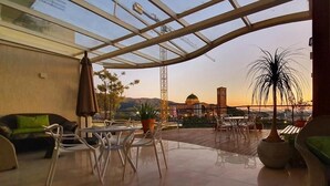 A spacious outdoor patio with a glass ceiling, a dining table, chairs, a sofa, and potted plants.