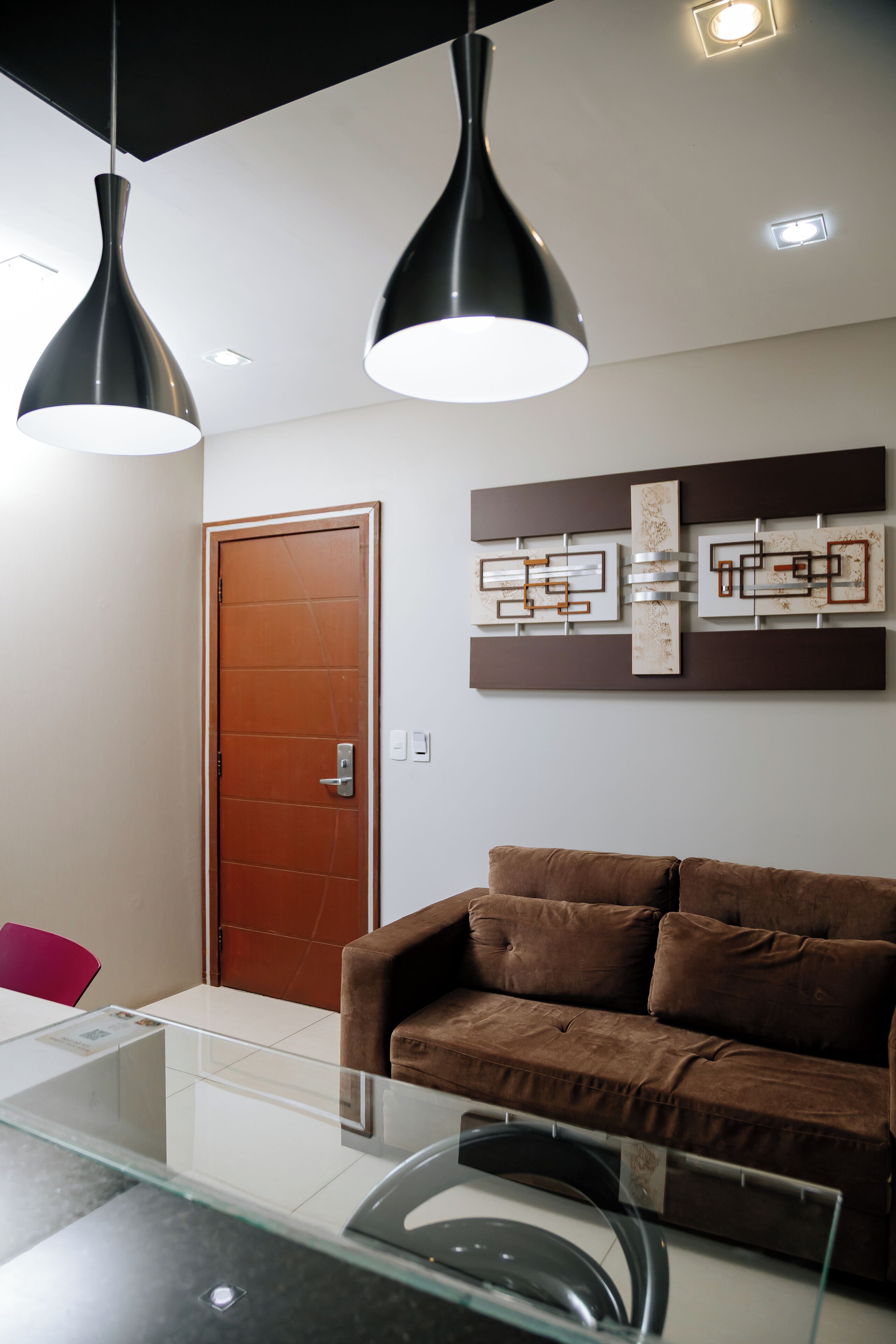 Uma sala de estar moderna com um sofá marrom, uma mesa de centro de vidro, luminárias pendentes e uma obra de arte abstrata na parede.