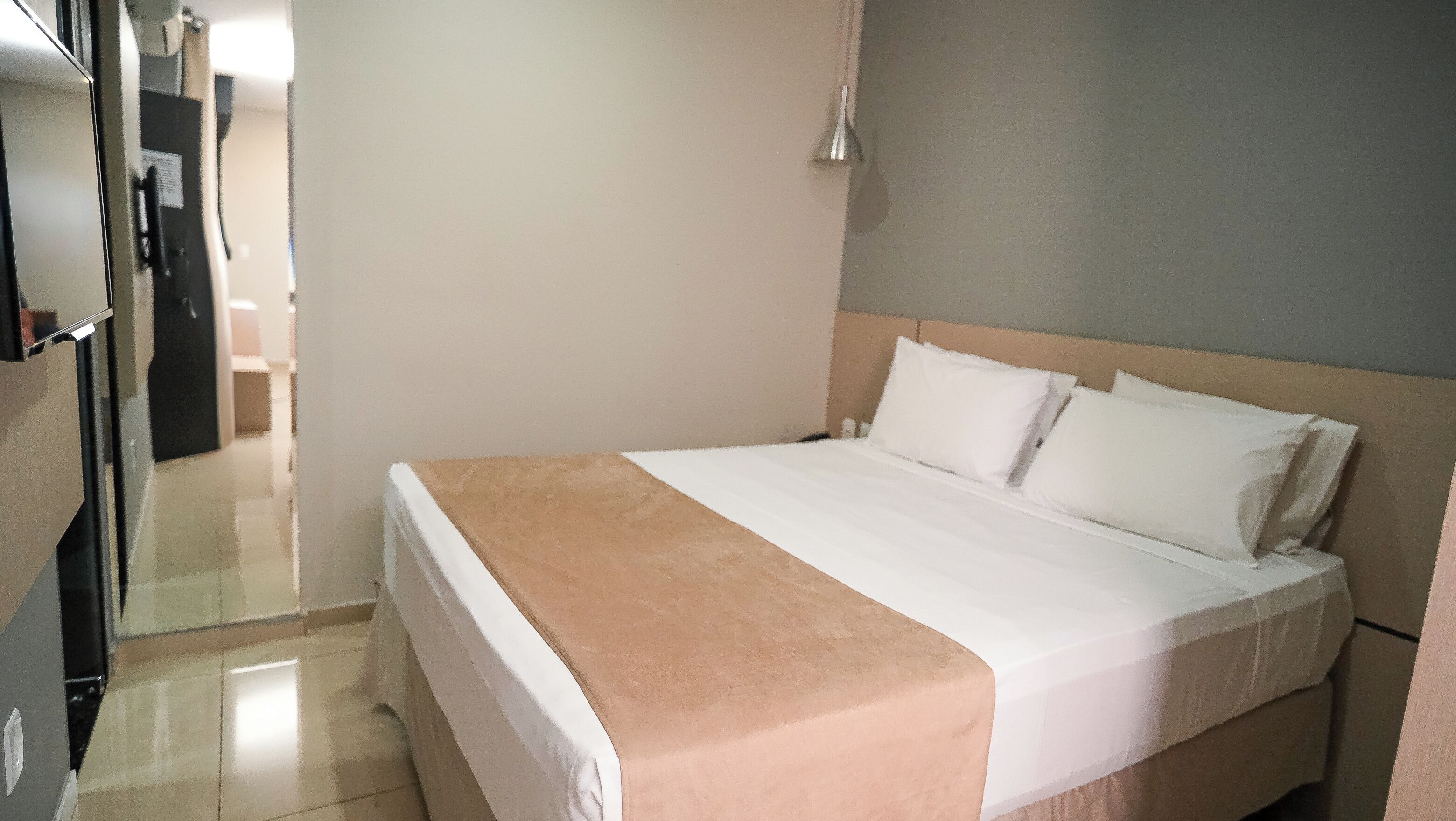 Quarto de hotel com cama, mesa de cabeceira, abajur e porta para um corredor.